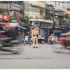 Artistic colour photo print of a street scene in Hanoi, Vietnam depicting movement blur of passing traffic and a still traffic warden in the middle.